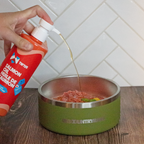 Person pouring Tuna brand oil into a green pet bowl with food on a wooden surface.
